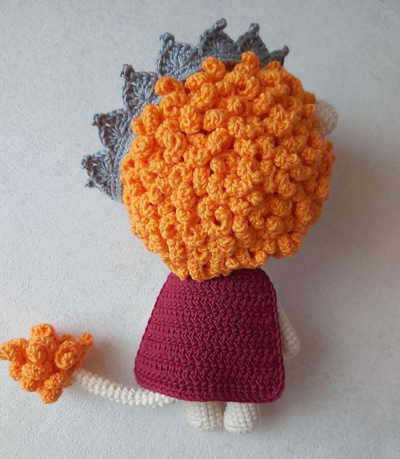 May include: A crocheted lion toy with a gray crown, orange mane, burgundy cape, and a white tail.