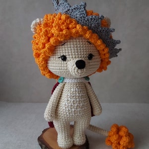 May include: A crocheted lion toy wearing a crown. The lion is white with orange hair and a tail. The crown is gray with orange accents.