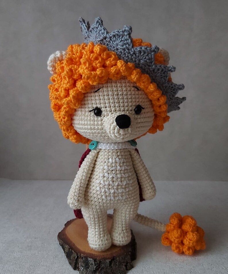 May include: A crocheted lion toy wearing a crown. The lion is white with orange hair and a tail. The crown is gray with orange accents.