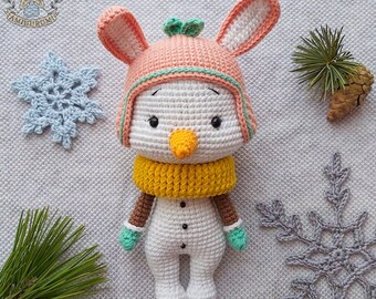 May include: A crocheted snowman amigurumi toy wearing a pink hat with bunny ears, a yellow scarf, and green mittens. The snowman is standing on a white textured surface with two gray snowflakes.