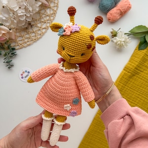 May include: A crocheted giraffe doll wearing a pink dress with flower accents. The doll has a yellow body, brown spots, and a white face. The doll is being held by a person's hands.