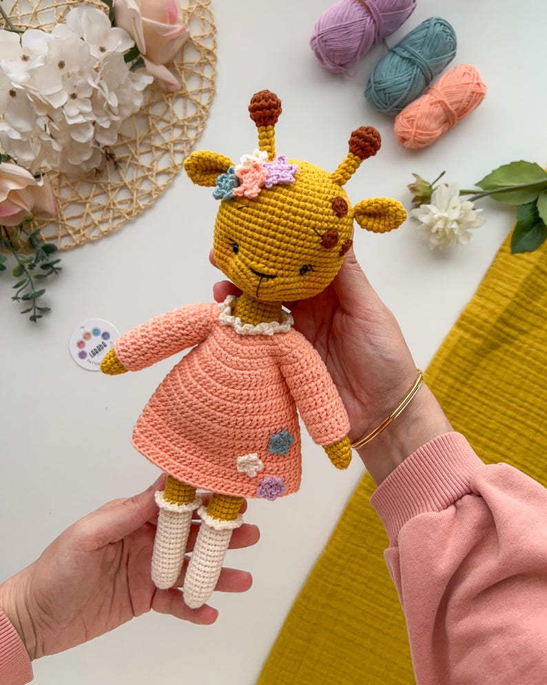 May include: A crocheted giraffe doll wearing a pink dress with flower accents. The doll has a yellow body, brown spots, and a white face. The doll is being held by a person's hands.