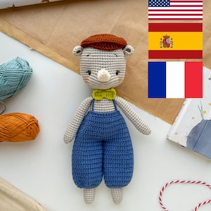 Può includere: Un peluche di stoffa a uncinetto a forma di rinoceronte che indossa un cappello marrone, una salopette blu e un papillon giallo. Il rinoceronte è in piedi su una superficie bianca.