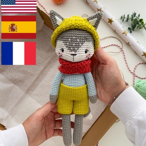 May include: A crocheted gray wolf toy wearing a yellow hat, red scarf, and yellow shorts. The toy is being held by a person's hands.