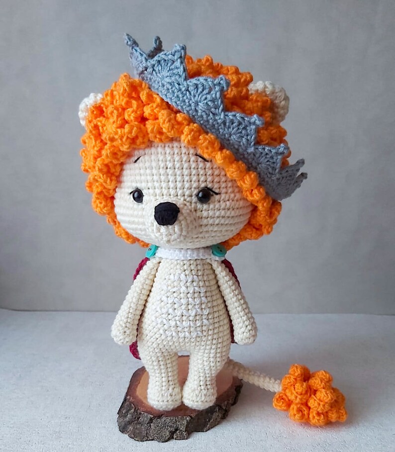 May include: A crocheted lion toy wearing a gray crown and a red cape. The lion has a white body, orange mane and tail, and black eyes.