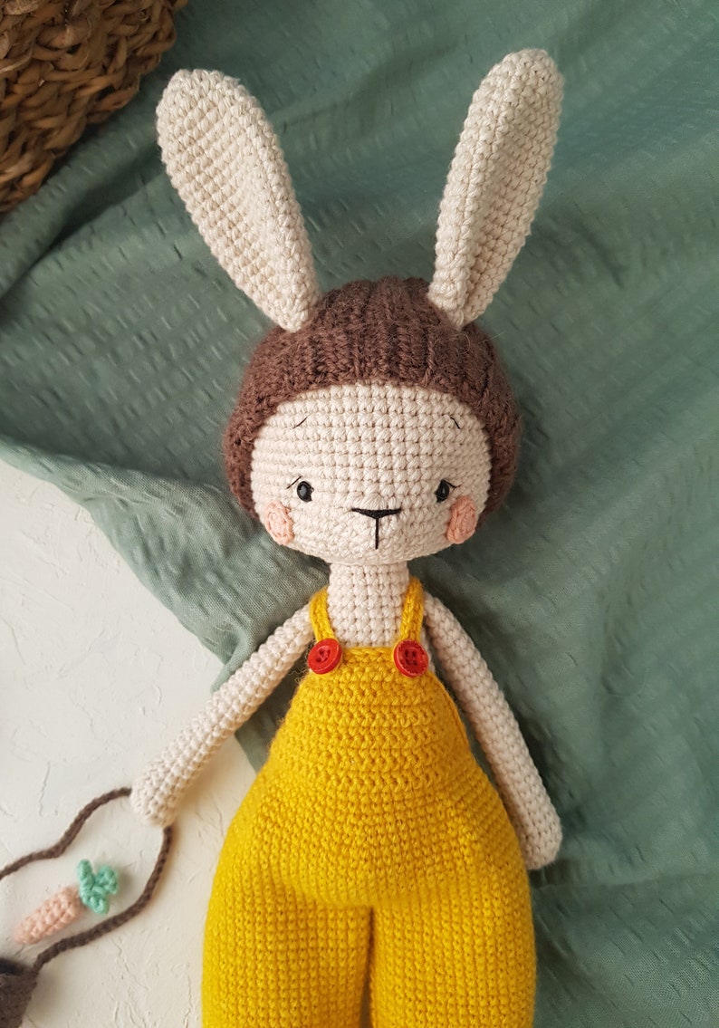 May include: A handmade crochet bunny doll with a cream-colored face, ears, and limbs. The bunny wears a brown cap and a yellow jumpsuit with red buttons. It holds a small bag with a carrot.