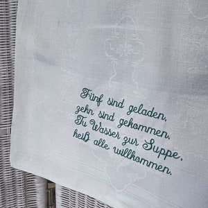 May include: White linen kitchen towel with green embroidered text that reads "F&uuml;nf sind geladen, zehn sind gekommen. Tu Wasser zur Suppe, hei&szlig; alle willkommen."