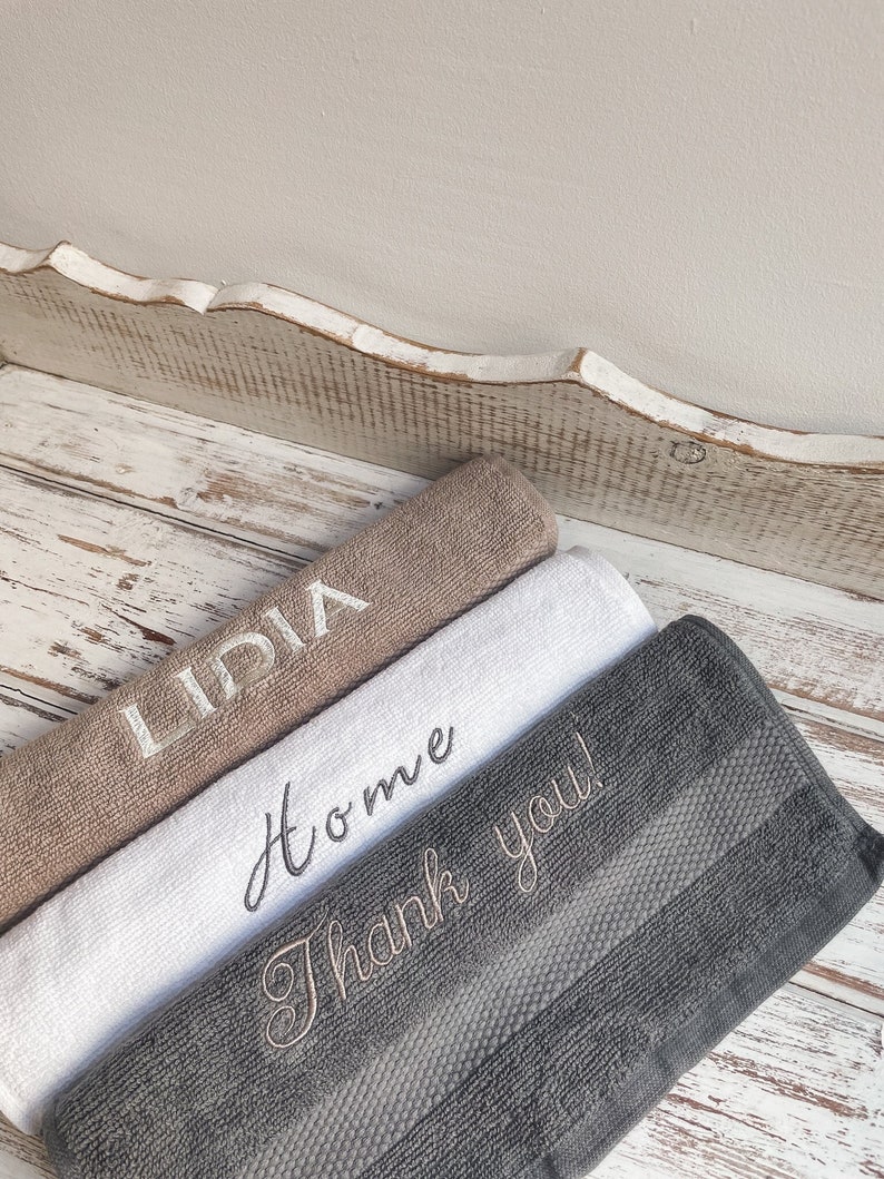 May include: Three towels stacked on a white wooden surface. The top towel is brown with the word "INDIA" embroidered in white. The middle towel is white with the word "Home" embroidered in black. The bottom towel is gray with the words "Thank you!" embroidered in brown.