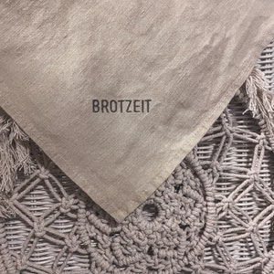 Puede incluir: Un mantel de lino beige con la palabra "BROTZZEIT" bordada en hilo negro. El mantel est&aacute; drapeado sobre una cesta de mimbre tejida.