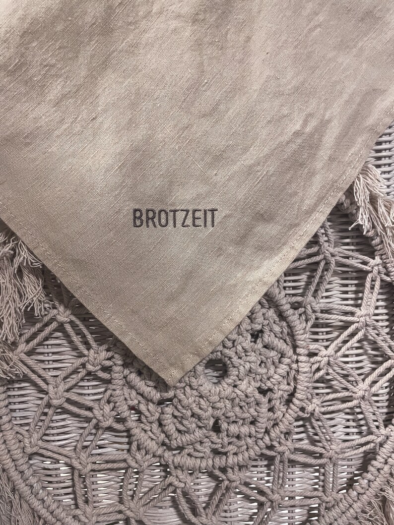 Puede incluir: Un mantel de lino beige con la palabra "BROTZZEIT" bordada en hilo negro. El mantel est&aacute; drapeado sobre una cesta de mimbre tejida.