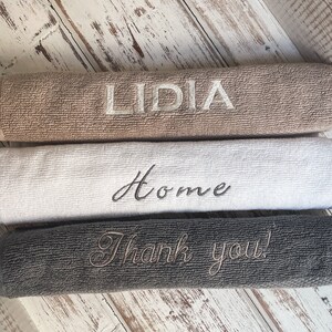 May include: Three towels stacked on a white wooden surface. The top towel is brown with the name "LIDIA" embroidered in white. The middle towel is white with the word "Home" embroidered in black. The bottom towel is gray with the phrase "Thank you!" embroidered in white.