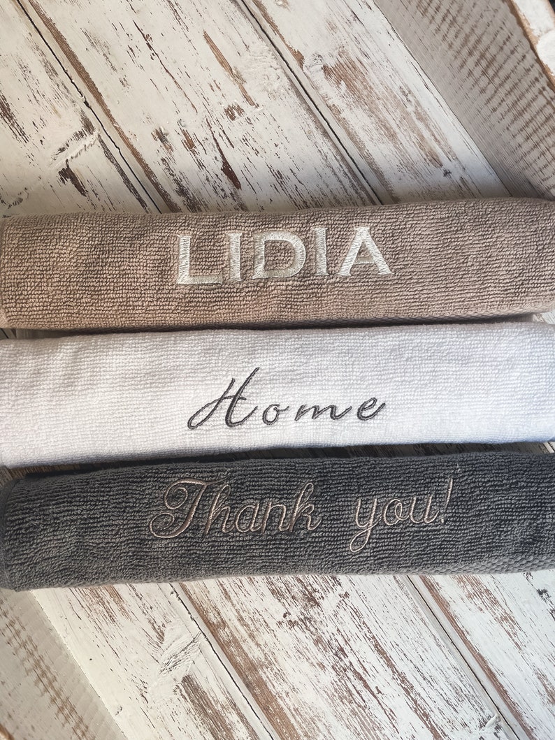 May include: Three towels stacked on a white wooden surface. The top towel is brown with the name "LIDIA" embroidered in white. The middle towel is white with the word "Home" embroidered in black. The bottom towel is gray with the phrase "Thank you!" embroidered in white.
