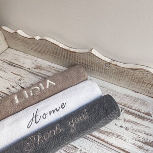 May include: Three towels stacked on a white wooden surface. The top towel is brown with the name "Lidia" embroidered in white. The middle towel is white with the word "Home" embroidered in black. The bottom towel is gray with the phrase "Thank you!" embroidered in white.