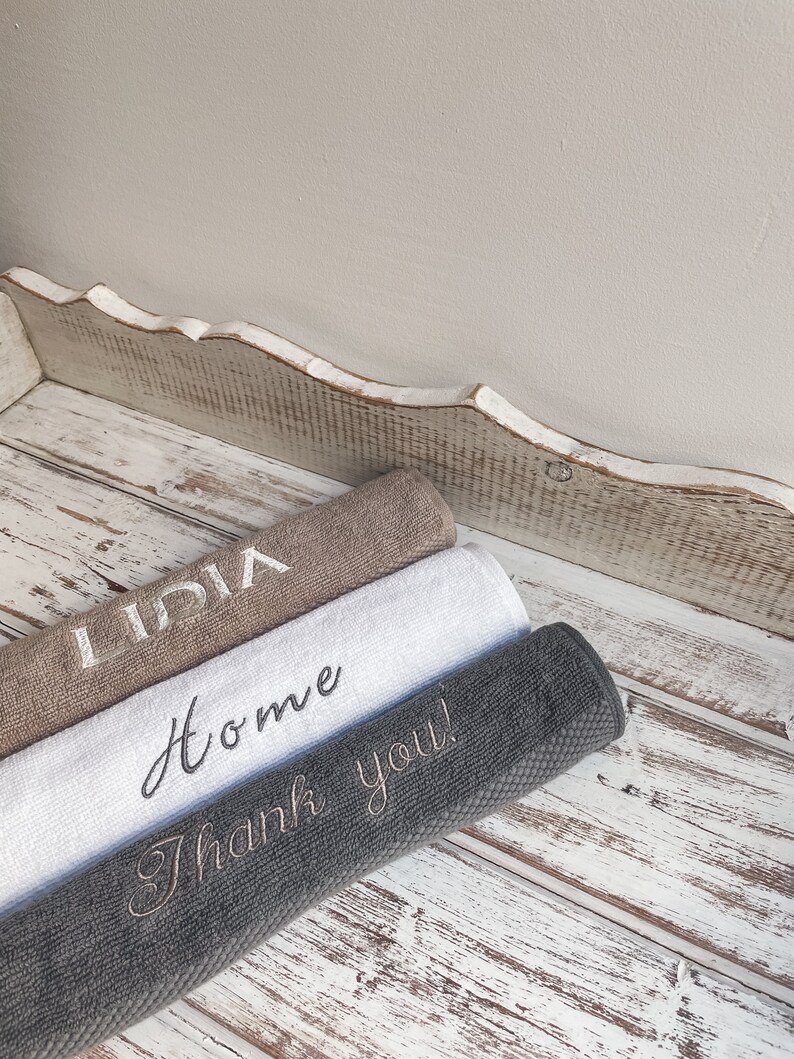 May include: Three towels stacked on a white wooden surface. The top towel is brown with the name "Lidia" embroidered in white. The middle towel is white with the word "Home" embroidered in black. The bottom towel is gray with the phrase "Thank you!" embroidered in white.
