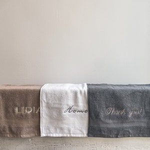 May include: Three towels with embroidered text. The towel on the left is brown with the word "LIDIA" embroidered in white. The middle towel is white with the word "Home" embroidered in black. The towel on the right is gray with the text "Thank you!" embroidered in white.