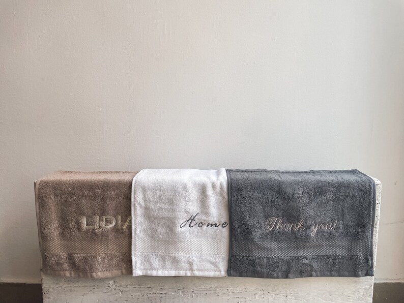 May include: Three towels with embroidered text. The towel on the left is brown with the word "LIDIA" embroidered in white. The middle towel is white with the word "Home" embroidered in black. The towel on the right is gray with the text "Thank you!" embroidered in white.