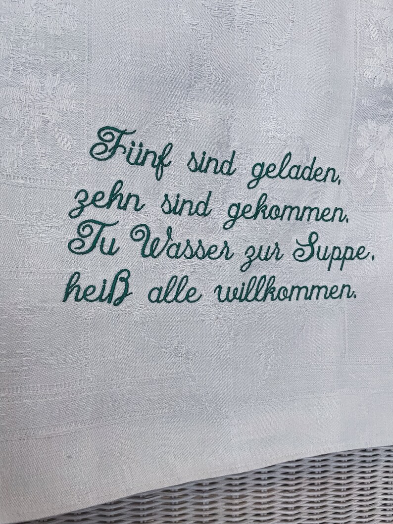 May include: Embroidered white linen kitchen towel with the text "F&uuml;nf sind geladen, zehn sind gekommen. Tu Wasser zur Suppe, hei&szlig; alle willkommen."