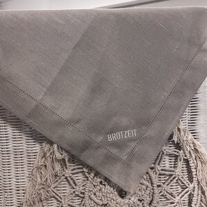 Puede incluir: Un camino de mesa de lino gris con un borde cosido y la palabra "BROTZET" bordada en blanco.