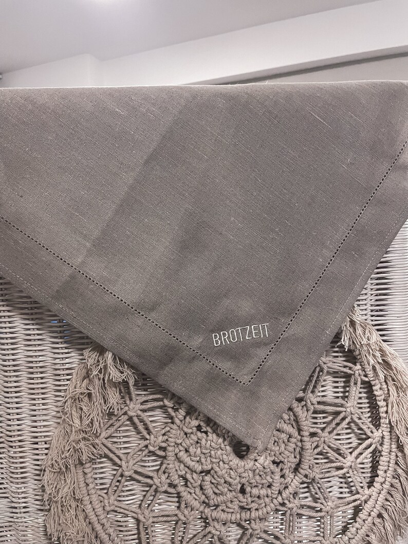 Puede incluir: Un camino de mesa de lino gris con un borde cosido y la palabra "BROTZET" bordada en blanco.