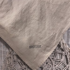 Puede incluir: Una bolsa de pan de lino beige con la palabra "BROTZEIT" impresa en negro.