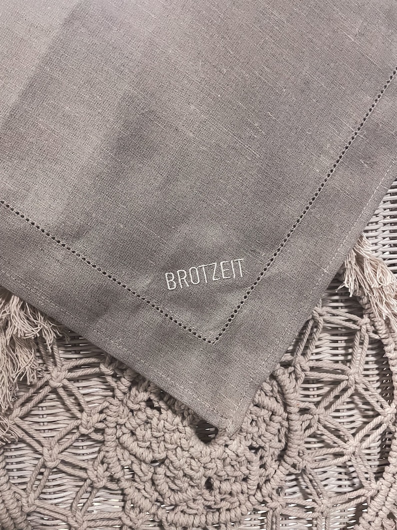 Puede incluir: Un camino de mesa de lino gris con un dise&ntilde;o bordado en blanco que dice "BROTZETIT". El camino de mesa est&aacute; doblado por la mitad y colocado sobre una superficie de mimbre tejido.