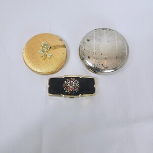 May include: Three vintage compacts. A round gold-toned compact with a floral embellishment, a silver-toned compact, and a rectangular black compact with a floral design in the center. The compacts are arranged on a white surface.