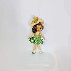 May include: A vintage doll with brown hair, wearing a cream-colored hat with a bow, a green and white patterned dress, and white shoes. The doll has painted facial features and is standing on a white surface.