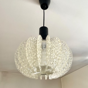 Könnte beinhalten: Eine klare Glaspendelleuchte mit schwarzem Kabel und Deckenbefestigung. Die Leuchte hat ein rundes, geschichtetes Design mit einer strukturierten Oberfläche.