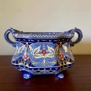 Peut inclure: Un pot de fleurs en céramique bleu de forme rectangulaire, orné de motifs floraux et de feuilles dorées, rouges et blanches. Il possède deux poignées incurvées et repose sur trois petits pieds. C'est un objet décoratif.