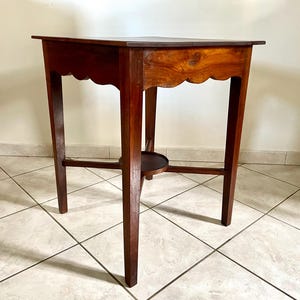 Peut inclure: Petite table en bois brun foncé avec une bordure festonnée et une petite étagère. La table a quatre pieds et mesure environ 71 cm de haut. Le grain du bois est visible sur le dessus.