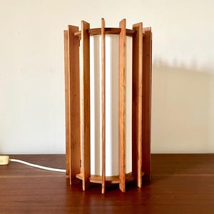 Może przedstawiać: Lampa stołowa w stylu mid-century modern o cylindrycznym kształcie. Lampa posiada pionowe drewniane listwy otaczające biały materiałowy klosz. Lampa ma około 38 cm wysokości i biały przewód zasilający.