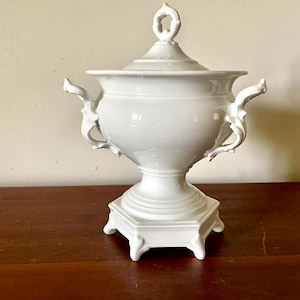 Puede incluir: Una urna blanca de cerámica con tapa, asas ornamentadas y base de pedestal. La urna tiene un adorno decorativo en la tapa y se asienta sobre una base cuadrada con pequeños pies. Pieza decorativa.