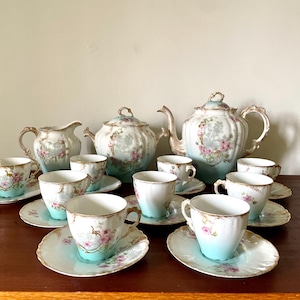 Peut inclure: Service à thé en porcelaine vintage comprenant une théière, un pot à crème, un sucrier et plusieurs tasses et soucoupes. L'ensemble présente un dégradé bleu clair avec des motifs floraux et des bordures dorées.