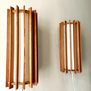 Może przedstawiać: Dwa drewniane kinkiety ścienne w stylu mid-century modern. Każdy kinkiet posiada pionowe drewniane listwy otaczające biały, cylindryczny dyfuzor światła. Kinkiety są prostokątne i mają ciepły, naturalny odcień drewna.