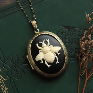 May include: A gold chain necklace with a black and white oval locket featuring a white ceramic bee. The locket is closed and has a detailed design.