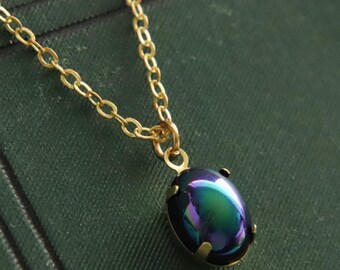 May include: A gold-colored chain necklace with a pendant featuring an iridescent, oval-shaped stone. The stone displays a blend of green, blue, and purple hues. The necklace is set against a dark green background, creating a striking contrast.
