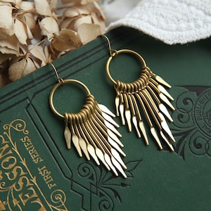 Puede incluir: Un par de pendientes dorados con una parte superior circular y formas alargadas en forma de lágrima colgantes. Los pendientes se muestran sobre un libro verde oscuro con letras doradas y flores secas en el fondo. Cierre de gancho.