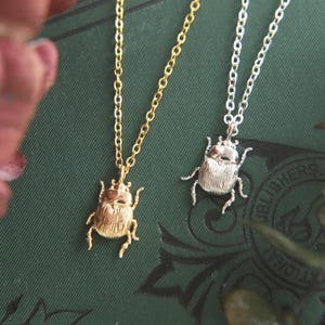 Puede incluir: Dos collares con colgantes de escarabajo, uno con acabado en tono dorado y otro en tono plateado. Cada colgante cuelga de una delicada cadena de metal a juego, mostrando texturas detalladas en el caparazón y las patas del insecto.