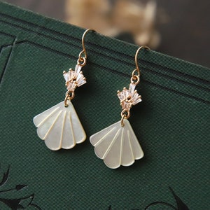 May include: A pair of gold earrings featuring a white shell-shaped pendant and a cluster of crystals. The earrings have a delicate and feminine design.