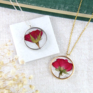 May include: Two delicate necklaces with round pendants featuring pressed red roses. The pendant on the left necklace has a silver frame, while the pendant on the right necklace has a gold frame.