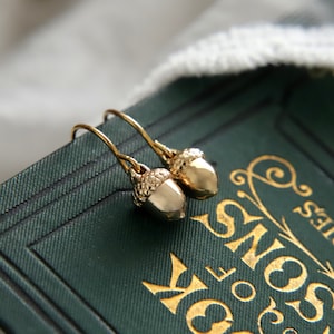 May include: A pair of gold-toned metal earrings shaped like acorns, featuring textured caps and smooth, polished bodies. Each earring hangs from a simple curved wire hook, showcasing a metallic finish that reflects light on the surface of the acorn.
