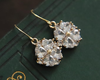 May include: A pair of gold-colored earrings with a floral design. Each earring features a cluster of clear, faceted stones arranged in a flower shape. The earrings have a simple hook closure.