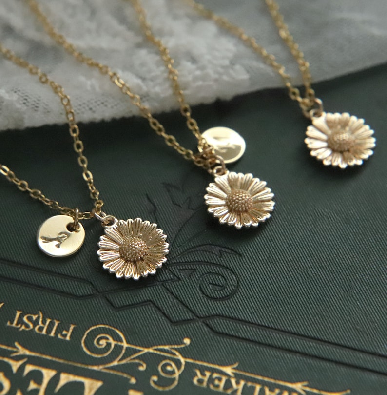 Puede incluir: Tres collares de oro con cadenas delicadas y colgantes. Cada colgante presenta un peque&ntilde;o disco de oro con una letra y un encanto de margarita de oro.