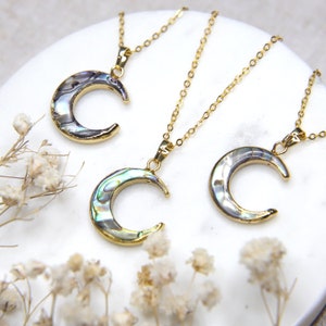 May include: Three gold necklaces with iridescent abalone shell crescent moon pendants. The necklaces are on a white surface.