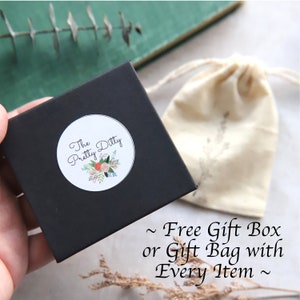 Puede incluir: Una caja de regalo negra con una pegatina blanca que dice "The Pretty Ditty" con un dise&ntilde;o floral. La caja est&aacute; sobre una superficie marr&oacute;n junto a una bolsa de tela blanca con cord&oacute;n. El texto "~ Free Gift Box or Gift Bag with Every Item ~" es visible debajo de la caja.