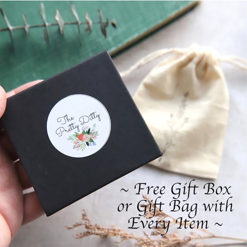 Puede incluir: Una caja de regalo negra con una pegatina blanca que dice "The Pretty Ditty" con un dise&ntilde;o floral. La caja est&aacute; sobre una superficie marr&oacute;n junto a una bolsa de tela blanca con cord&oacute;n. El texto "~ Free Gift Box or Gift Bag with Every Item ~" es visible debajo de la caja.