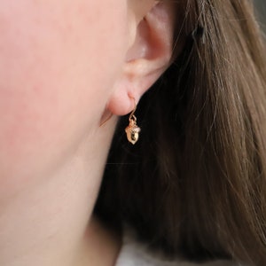 May include: A small gold-toned acorn charm earring featuring a textured cap and a smooth, pointed base. The charm hangs from a simple gold-toned hook wire, creating a delicate and minimalist piece of jewelry that rests against the earlobe.