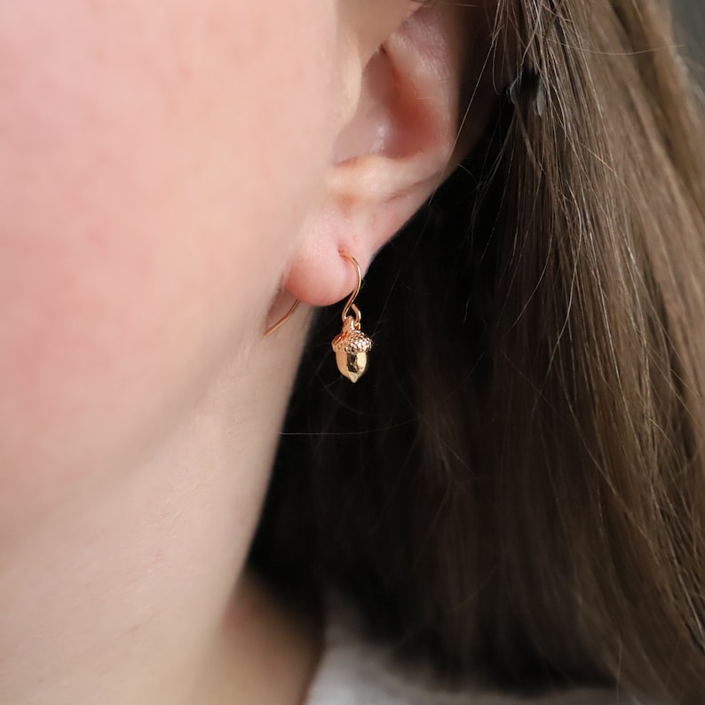 May include: A small gold-toned acorn charm earring featuring a textured cap and a smooth, pointed base. The charm hangs from a simple gold-toned hook wire, creating a delicate and minimalist piece of jewelry that rests against the earlobe.