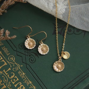 Puede incluir: Conjunto de collar y pendientes de oro con dise&ntilde;o de margarita. El collar tiene un peque&ntilde;o dije inicial y los pendientes son de estilo colgante.