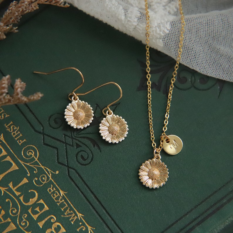 Puede incluir: Conjunto de collar y pendientes de oro con dise&ntilde;o de margarita. El collar tiene un peque&ntilde;o dije inicial y los pendientes son de estilo colgante.